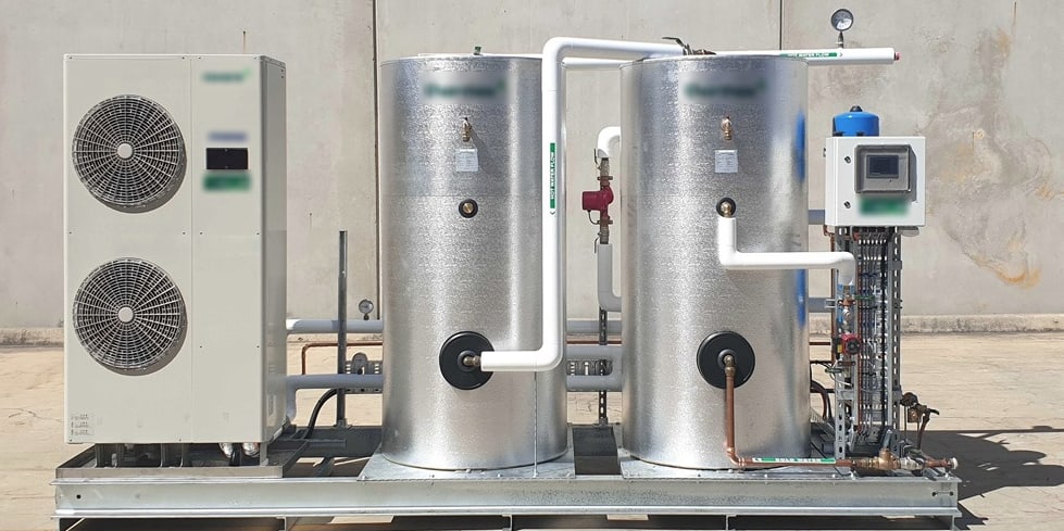 Skid-mounted commercial heat pump water heating system: an air source heat pump alongside two insulated storage tanks, connected by insulated flow and return pipework with a pump, valve set and controller.
