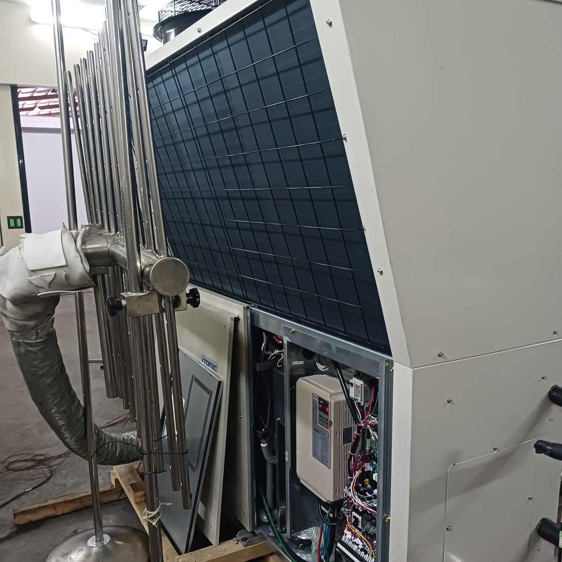 A commercial air source heat pump on a pallet with its electrical panel open, showing the inverter drive and wiring, with an air temperature measurement grid set up in front of the evaporator coil.
