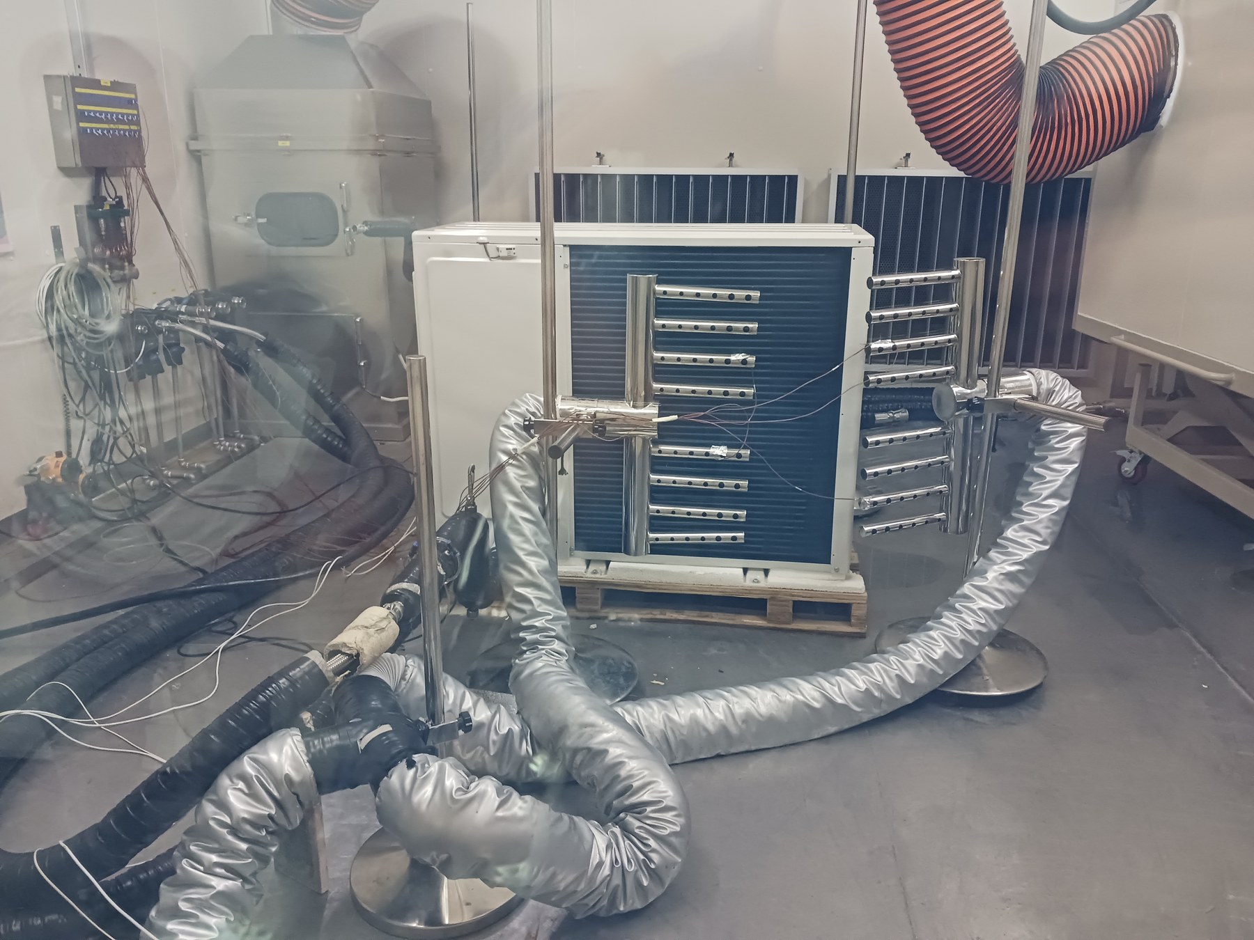 Outdoor-air side of a heat pump unit in a test chamber, with temperature sensors on stands and insulated ducts